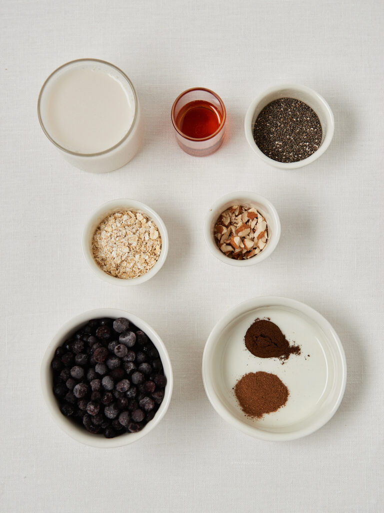 Flatlay of smoothie ingredients including berries, seeds, spices and liquids in small white bowls