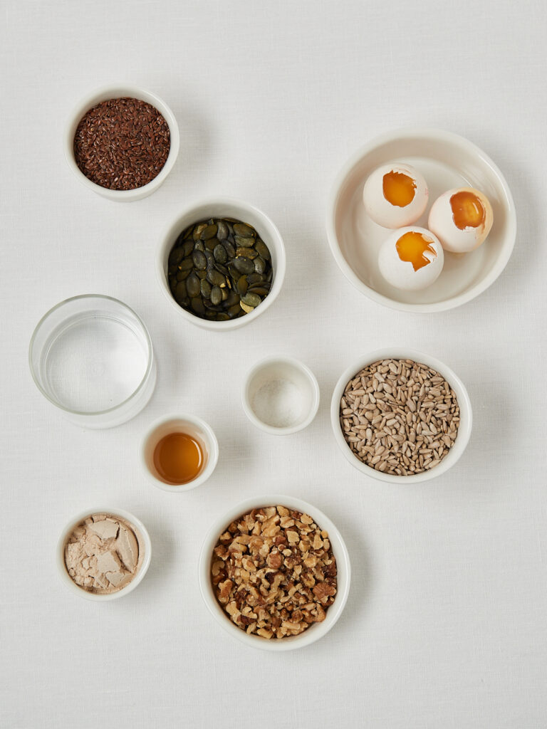 Zutaten für das Life-Changing-Bread in einzelnen Schälchen arrangiert: Leinsamen, Kürbiskerne, Sonnenblumenkerne, Nüsse, Haferflocken, Wasser, Ahornsirup und Eierschalen.