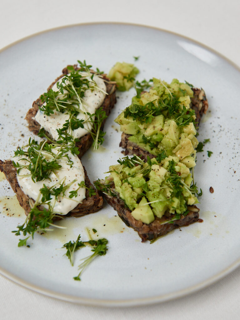 Avocado-Toast auf dunklem Brot, belegt mit cremiger Avocado, frischen Kräutern und Gewürzen, auf einem weißen Teller serviert.