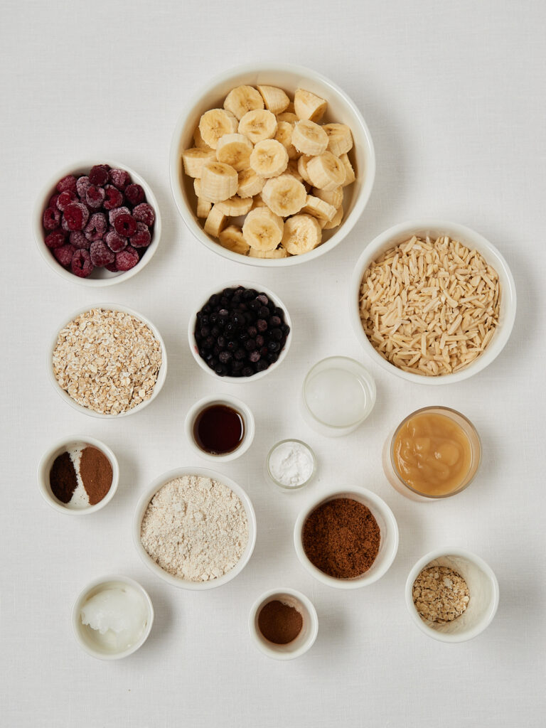 Flatlay of banana bread ingredients including bananas, oats, nuts, berries, spices and sweeteners in small bowls.