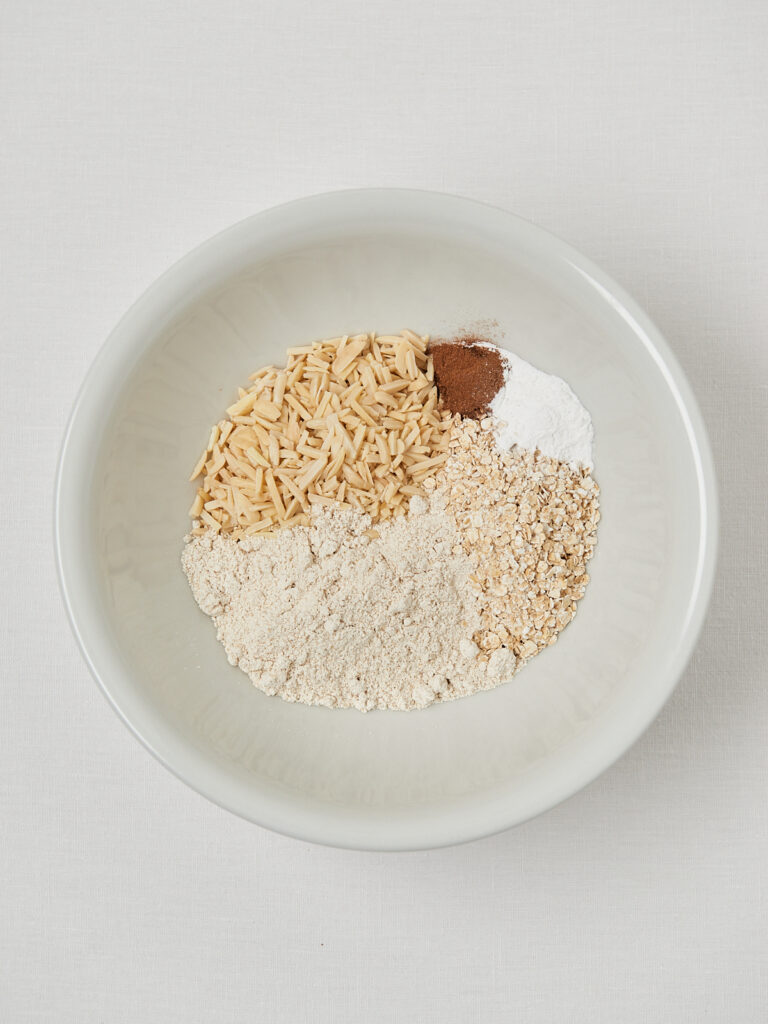 Oats, almond flour, baking powder, cinnamon and sliced almonds measured into a white bowl.