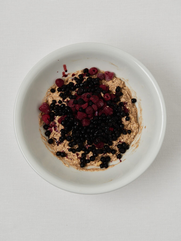 Frozen mixed berries added to banana bread batter before stirring, shown in a white bowl.
