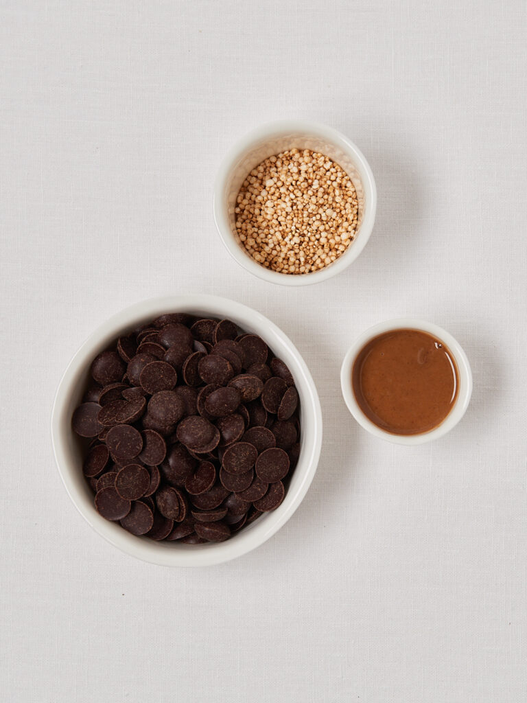 Flatlay of chocolate chips, puffed quinoa and creamy hazelnut butter in white bowls for hazelnut chocolate bites.