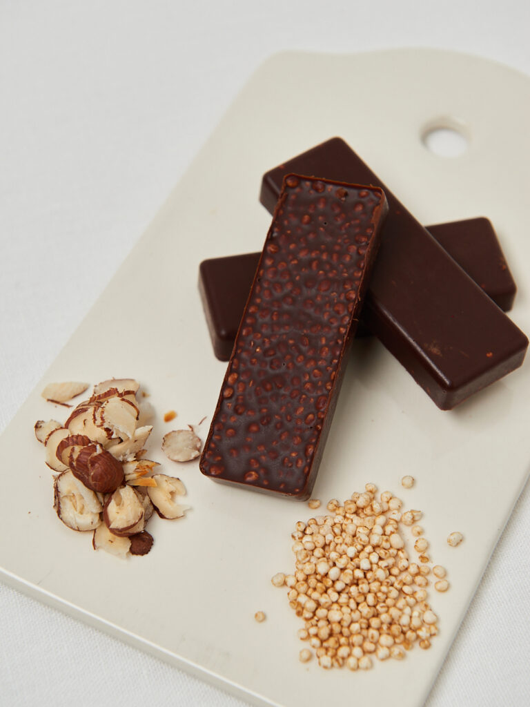 Fertige Schoko-Haselnuss-Riegel mit gepufftem Quinoa, angerichtet auf einem weißen Brett mit Haselnussstücken und Quinoa.