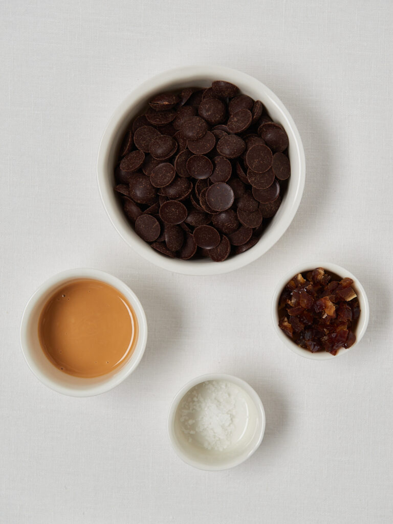 Zutaten f&uuml;r Schoko-Haselnuss-Bites: dunkle Schokodrops, Haselnussmus, gepuffter Quinoa und Salz in kleinen Sch&auml;lchen auf wei&szlig;em Untergrund.