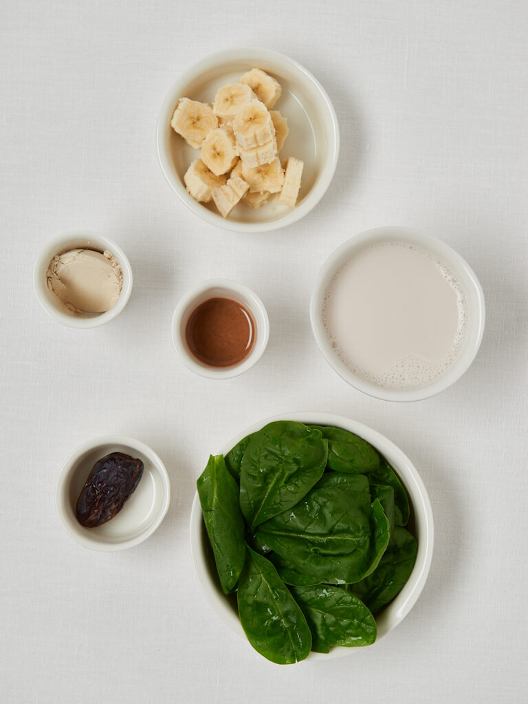 Zutaten f&uuml;r einen gr&uuml;nen Smoothie in kleinen Schalen: Bananenscheiben, Spinat, Pflanzenmilch, Nussmus, Ahornsirup und eine Dattel