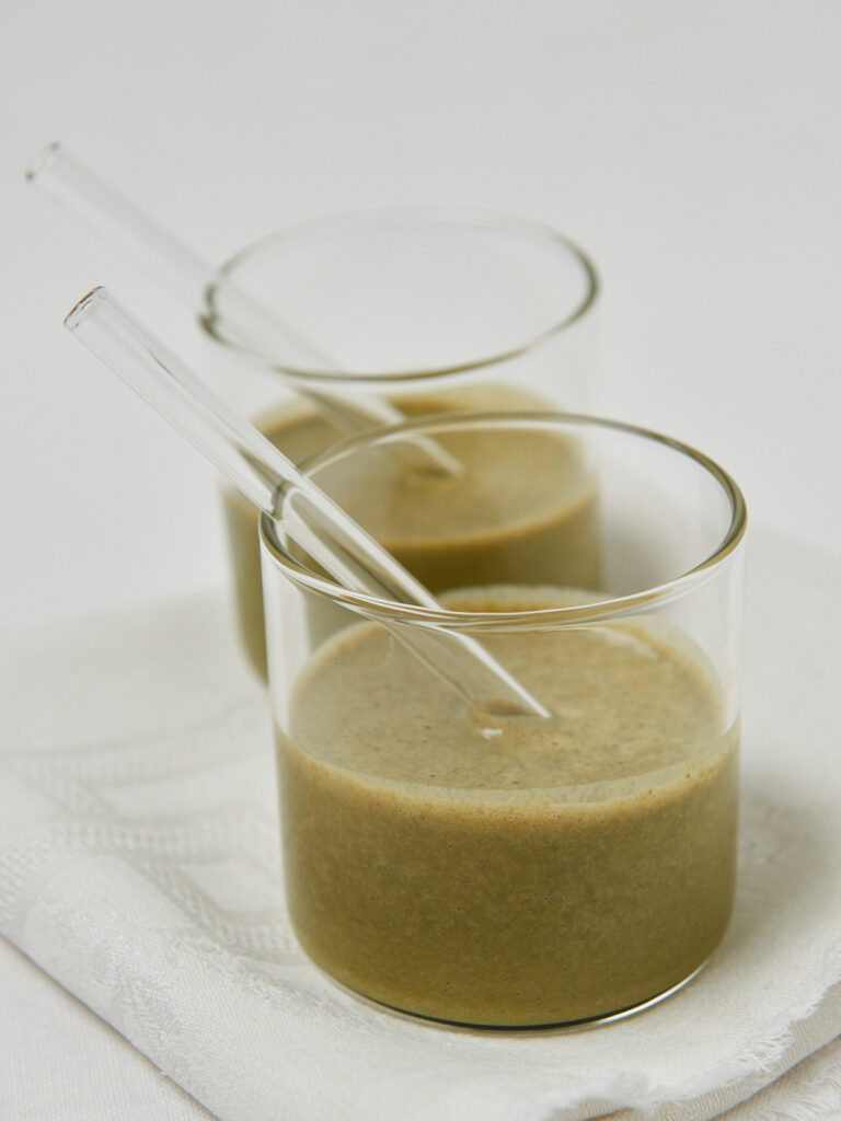 Zwei Gl&auml;ser mit gr&uuml;nem Smoothie und Glasstrohhalmen auf einem wei&szlig;en Tuch.