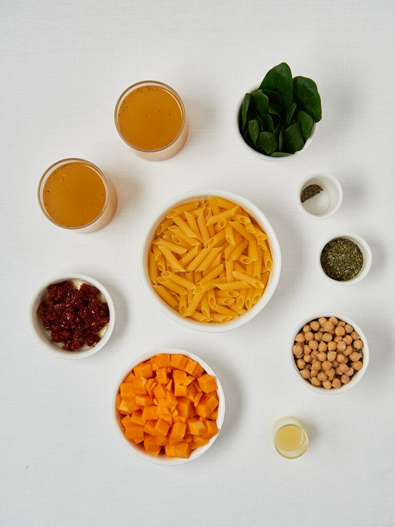 Flatlay der Zutaten f&uuml;r One-Pan-Pasta: Pasta, Kichererbsen, K&uuml;rbis, Spinat und Gew&uuml;rze