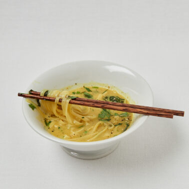 Vegane Ramen Nudeln in goldener Brühe mit Kräutern auf hellem Hintergrund