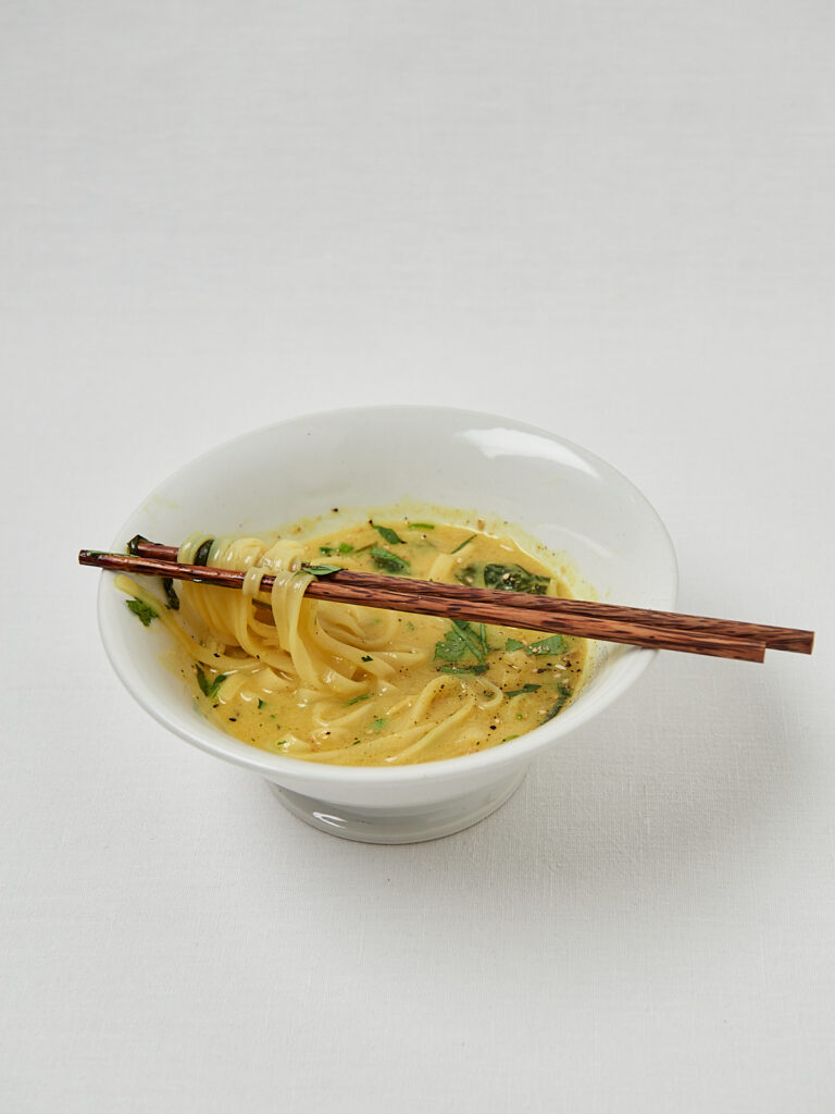 Vegane Ramen Nudeln in goldener Br&uuml;he mit Kr&auml;utern auf hellem Hintergrund