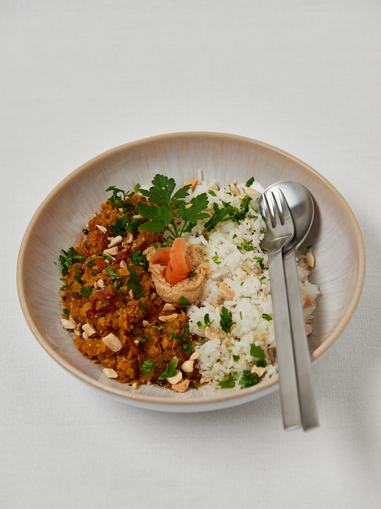 Rotes Linsen Dal mit Koriander und Cashews in einer Keramikschale, daneben ein Dessertlöffel und eine Gabel