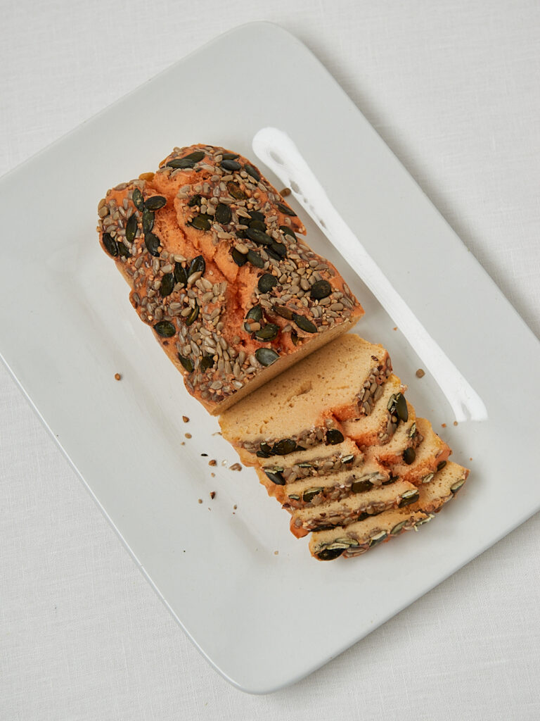 Aufgeschnittenes Linsenbrot mit K&uuml;rbiskernen und Karotten auf wei&szlig;em Schneidebrett &ndash; seitliche Aufnahme