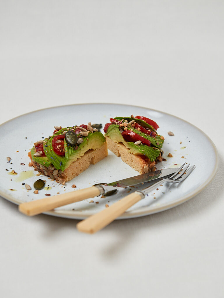 Linsenbrot Scheibe mit Avocado, Chiliflocken und K&uuml;rbiskernen auf wei&szlig;em Teller mit Besteck